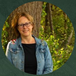 Portretfoto van Grietje Hofsteenge, een vrouw met halflang krullend haar en een bril, gekleed in een spijkerjack en zwart shirt, met een groene, bosrijke achtergrond.
