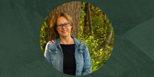 Portretfoto van Grietje Hofsteenge, een vrouw met halflang krullend haar en een bril, gekleed in een spijkerjack en zwart shirt, met een groene, bosrijke achtergrond.