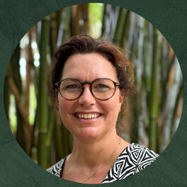 Portretfoto van Caroline Bardoel, een vrouw met kort bruin haar en een bril, lachend, gekleed in een zwart-wit patroon blouse, met een achtergrond van bamboe.