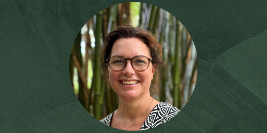 Portretfoto van Caroline Bardoel, een vrouw met kort bruin haar en een bril, lachend, gekleed in een zwart-wit patroon blouse, met een achtergrond van bamboe.